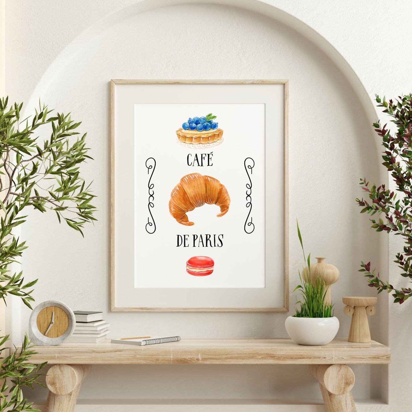 Framed artwork with a croissant, macaron, and cake illustration on a wall above a wooden shelf.