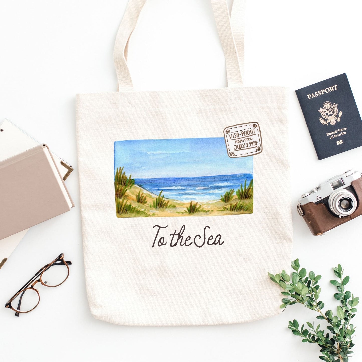 Tote bag with beach scene and 'To the Sea' text, surrounded by travel items on a white background